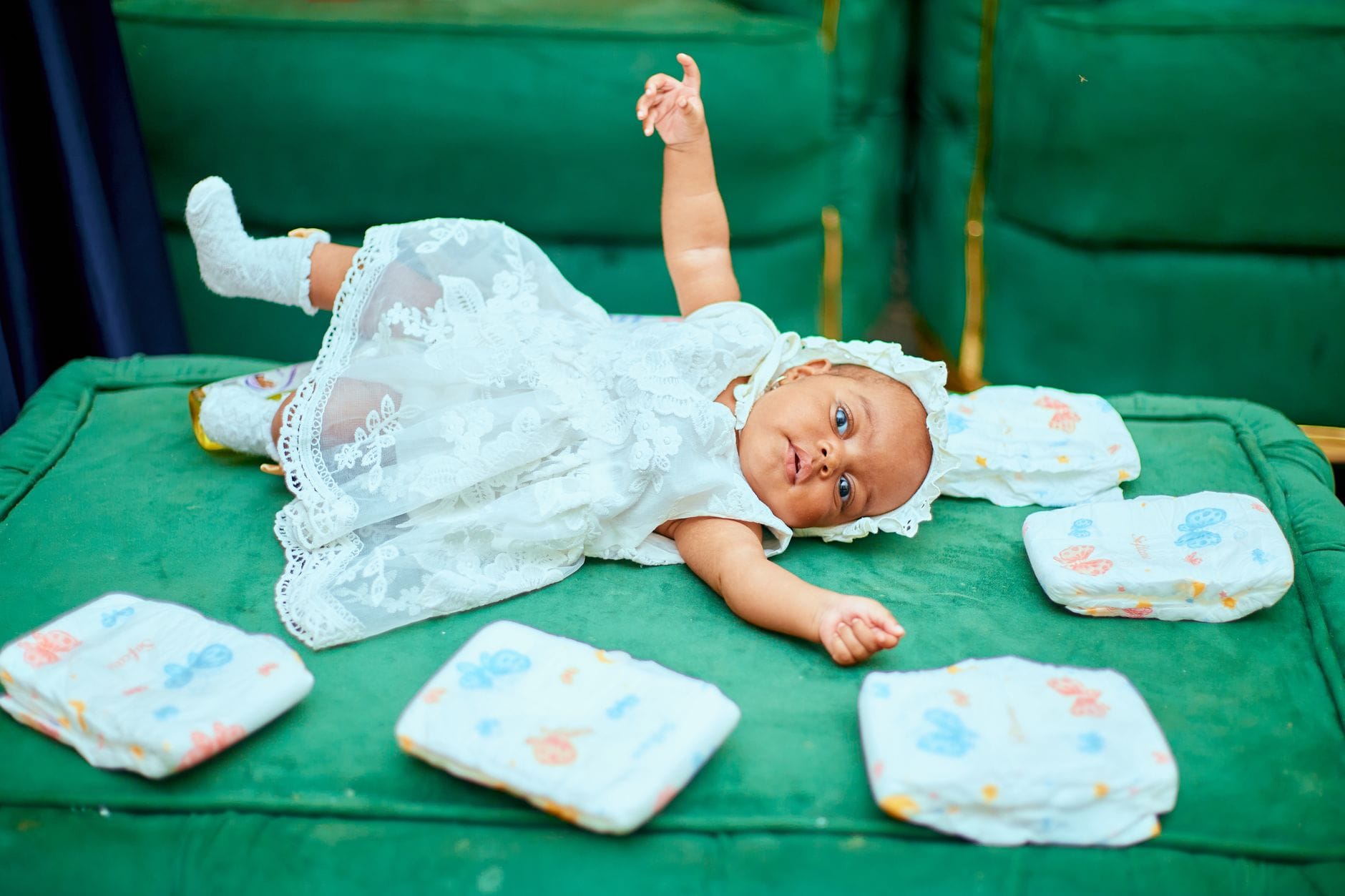 Niemowlę w białej sukience bawi się na zielonej sofie obok pieluszek